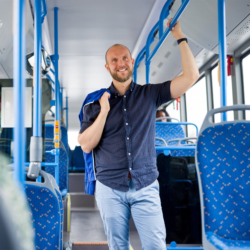 Mies seisoo bussissa, hymyilee ja pitää tangosta kiinni. 