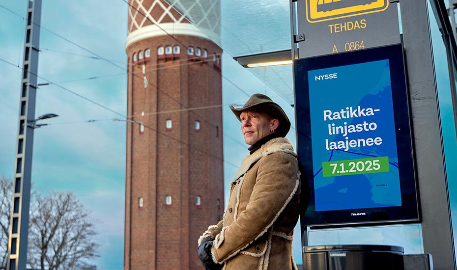 Cowboy-tyyliin pukeutunut mies nojaa Tehtaan ratikkapysäkkiä vasten Hiedanrannassa. 