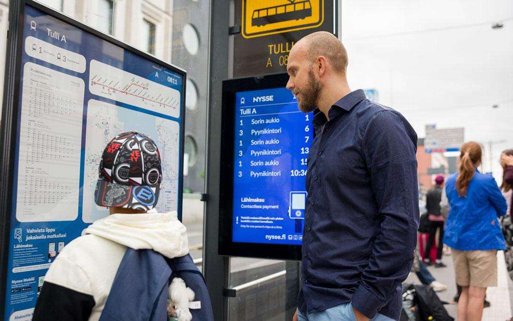 Lippalakkipäinen lapsi katselee vanhemman kanssa linjakarttaa ratikkapysäkillä. Taustalla näkyy digitaalinen aikataulunäyttö sekä muita matkustajia. Ilma on pilvinen.